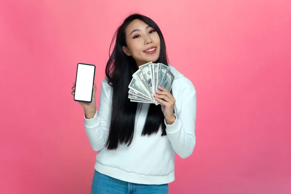 Šťastná mladá asijská dívka drží peníze a telefon s prázdnou obrazovku v rukou. Bankovní nebo online kasino aplikace — Stock fotografie
