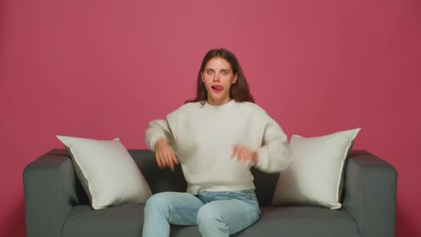 Zabawna zabawna młoda dziewczyna sprawia, że twarze wybrzuszone uszy pokazują język, dokuczanie, wygłupy, siedzenie na kanapie na różowym tle — Wideo stockowe