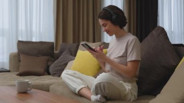 Modern genç kadın kulaklıkla favori müzik dinliyor cep telefonu müzik uygulaması kullanıyor, evdeki kanepede oturuyor. Yüksek kalite 4k görüntü