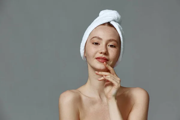 Όμορφη νεαρή κοπέλα με πετσέτα στο κεφάλι, καθαρό υγιές δέρμα μετά την περιποίηση της επιδερμίδας σε γκρι φόντο στούντιο — Φωτογραφία Αρχείου
