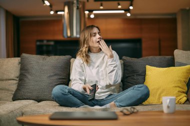 Sıkıcı haberlerden, yayınlardan, evdeki kanepede oturmaktan bıkmış, elinde uzaktan kumandayla esneyen genç bir kadın.