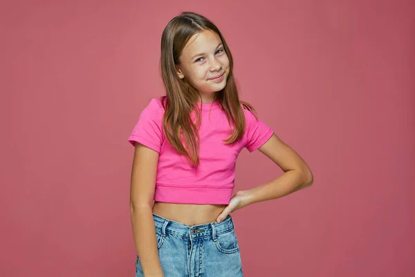 Linda niña sonriente moderna mirando a la cámara, posando sobre fondo de estudio rosa. Concepto de infancia feliz —  Fotos de Stock