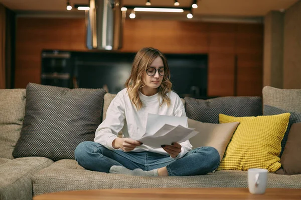 Gözlüklü, ciddi genç bir bayan kanepede oturuyor. Evrak, fatura okuyor, ev işleriyle uğraşıyor. — Stok fotoğraf