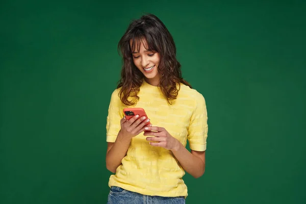 Smiling modern young girl hold phone looking at smartphone using mobile dating apps, chatting online on green background Stock Picture