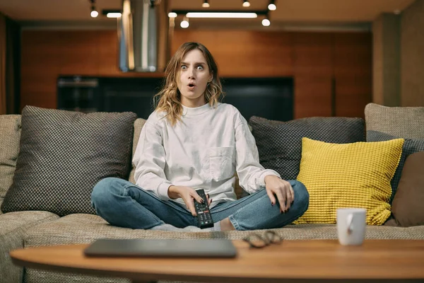 Sorprendida joven mujer viendo seriales o películas, sentada en el sofá, diciendo wow, maravillada por la inesperada trama de la película Fotos de stock libres de derechos