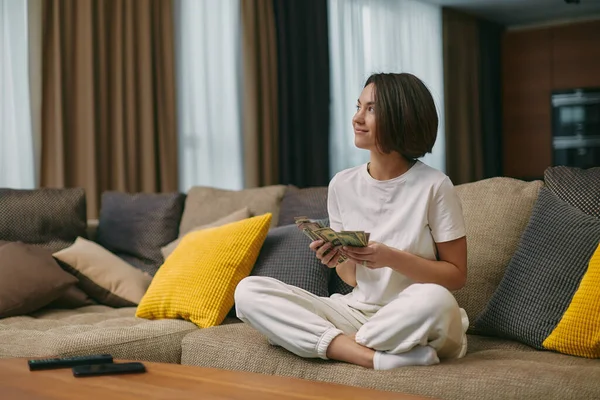 Young woman housewife holding money, dreaming about shopping, thinking how spend cash, sitting on sofa in living room Stock Picture