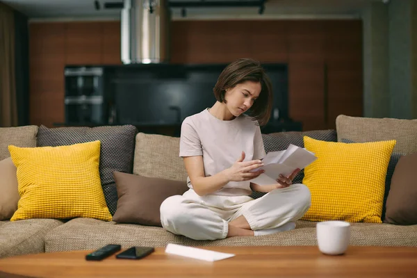 Mujer joven estresada mirando el documento en papel, sentada en el sofá en casa, recibió la factura del apartamento o del alquiler Imágenes de stock libres de derechos