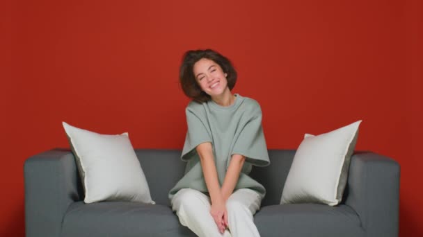赤い壁を持つ部屋のソファの上の美しい女の子は甘く微笑み、冗談で笑います。コメディとユーモラスなテレビ番組. — ストック動画