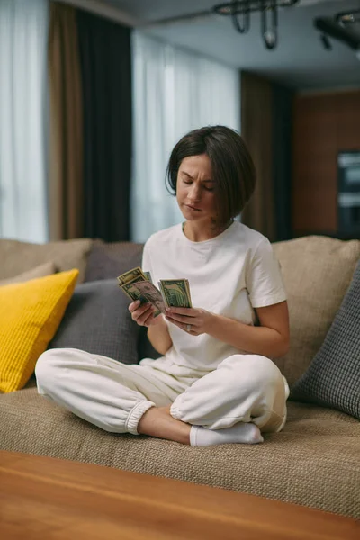Ciddi şekilde kaşlarını çatmış genç bir kadın para sayıyor, maddi sorunlar, borç, evdeki kanepede oturuyor. — Stok fotoğraf