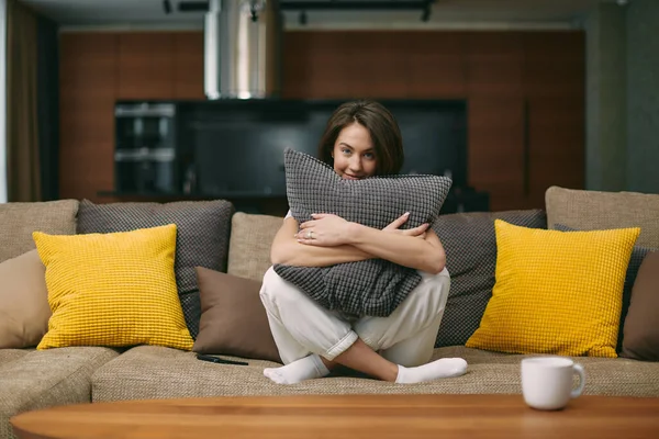 Piękna młoda kobieta przytula poduszkę siedząc na wygodnej sofie w salonie, odpoczywając w domu, czując spokój — Zdjęcie stockowe