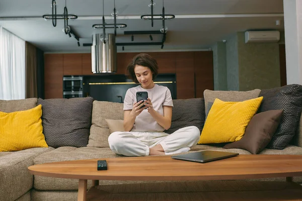 Evdeki koltukta oturan genç kız sosyal medya kuryesini kullanıyor, internette sörf yapıyor, internetten alışveriş yapıyor. — Stok fotoğraf
