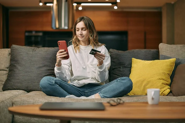 Młoda kobieta posiada bankową kartę kredytową, wpisuje informacje o płatnościach przez telefon, kupuje online siedząc na kanapie w domu Zdjęcie Stockowe