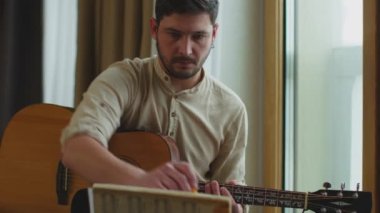 Yaratıcı genç müzisyen akustik gitar çalıyor, pencere pervazında oturan notalara bakıyor. Kodlayıcıyı Düzenle