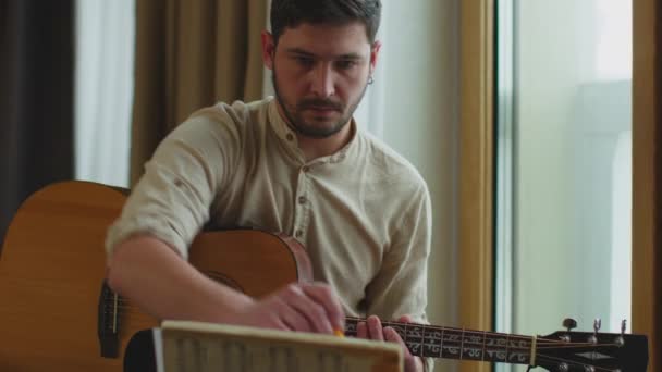 Creatief getalenteerde jonge man muzikant die akoestische gitaar speelt, kijkend naar bladmuziek zittend op vensterbank. Prores-codec — Stockvideo