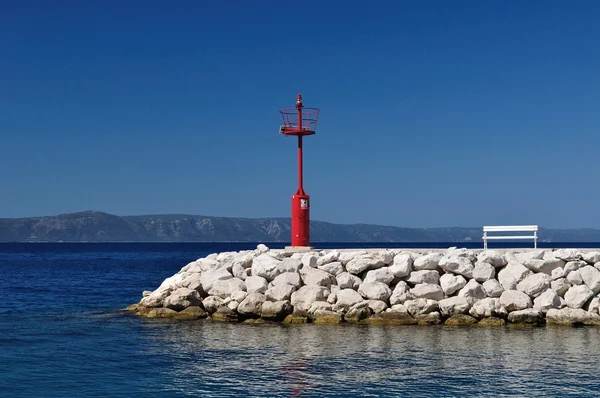 Красный маяк в порту с камнями и белой скамейкой — стоковое фото