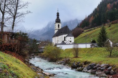 Ramsau, Germany - November 29, 2019: St. Sebastian church next to a creek with the Bavarian Alps in the background on a cloudy fall day.
