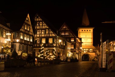 Gengenbach, Germany - December 13, 2020: Half-timbered buildings and clock tower lit up at night on a street with Christmas trees and decorations during lockdown in Gengenbach, Germany.