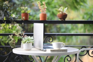 Gümüş dizüstü bilgisayar ve kahve fincanı, beyaz masada defterler ve balkonda saksılar.