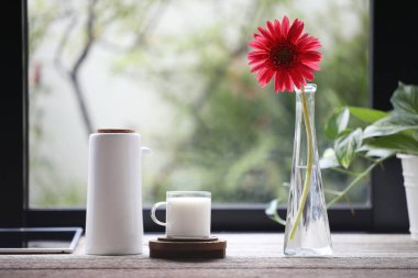 Bir bardak süt, sürahi sürahi ve cam vazoda çiçek pencerenin önündeki ahşap masada.
