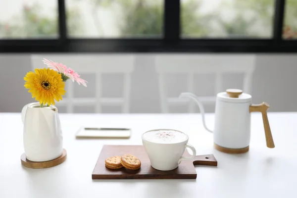 cappuccino coffee and sandwich cookie and gerbera flower on white wooden table