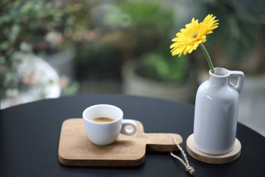 Siyah ahşap masa üzerinde sarı gerbera çiçekli beyaz bir fincanda kahve.