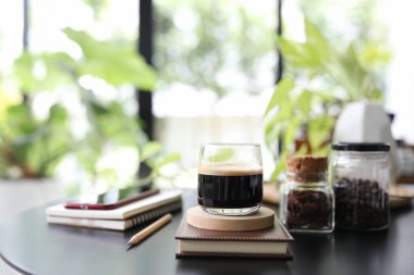 Cam bir fincanda Espresso kahvesi, defter, telefon ve siyah masaya demlik.