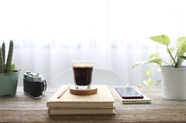 Cam bir fincanda kremalı kahve, defterler ve ahşap masada bitkilerle birlikte telefon.