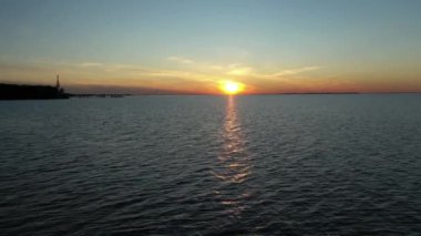 Gün batımında deniz ya da okyanus yüzeyinde uçan bir dronun hava görüntüsü, sudaki turuncu güneşin yansıması. Dalgalar ve su yüzeyinde parlayan ışık.