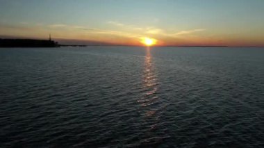 Gün batımında deniz ya da okyanus yüzeyinde uçan bir dronun hava görüntüsü, sudaki turuncu güneşin yansıması. Dalgalar ve su yüzeyinde parlayan ışık.