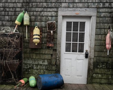 Bernard, ME - USA - Oct.15,2021: Horitzonal view of a wooden door to a shingled workshop in a dock area, flanked by lobster traps, trap buoys, rope and other fishing implements. clipart