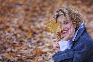 Genç güzel kız gülümsüyor ve elinde sarı bir akçaağaç yaprağı tutuyor. Kızın kıvırcık saçları var. Autumn Park 'ta. Uzayı kopyala Bulanık arkaplan.