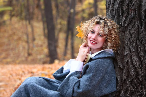Молодая девушка с вьющимися волосами в сером пальто в осеннем парке. Девушка сидит под деревом с книгой в руках. Время года - осень. Желтые листья на земле. Листопад в парке. — стоковое фото