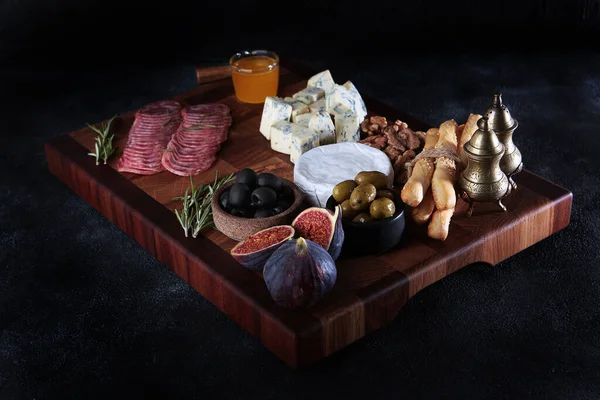 Several types of cheese on a wooden board with smoked sausage and olives. Figs and grissini. Honey and cheese on wooden board. Selective focus. Dark background. — Stock Photo, Image