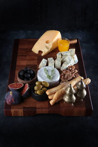 Several types of cheese on a wooden board with smoked sausage and olives. Figs and grissini. Honey and cheese on wooden board. Selective focus. Dark background. Vertical photo. Royalty Free Stock Photos