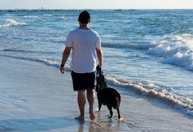 Meksikalı bir adam köpeğiyle Meksika 'da bir Riviera Maya plajında yürüyor. Arka plan görüntüsü. Arka planda Karayip Denizi ve demirli tekneler var..