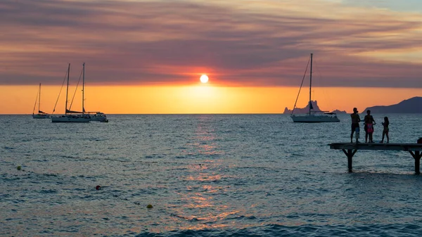 Three friends on the pier watch the sunset over the sea and sailboats anchored on the island of Formentera in Spain