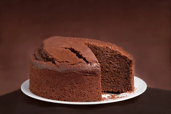 A round chocolate sponge cake with some missing slices on a white plate. Brown background.