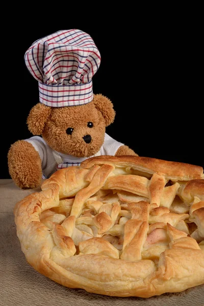 A Teddy bear dressed as chef cook with a chef hat and an apron is showing off his freshly baked apple pie. Black background.