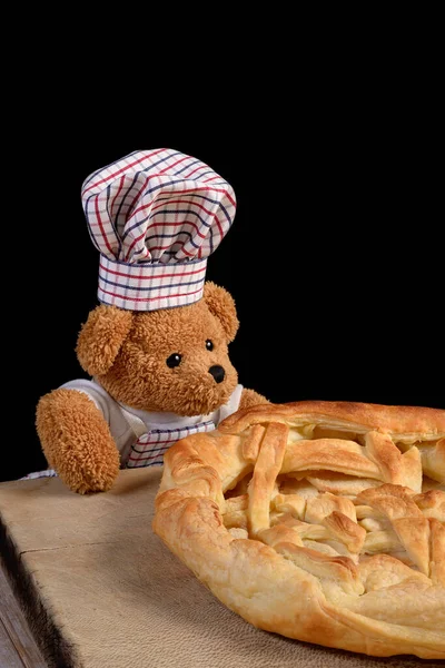 A Teddy bear dressed as chef cook with a chef hat and an apron is showing off his freshly baked apple pie. Black background.