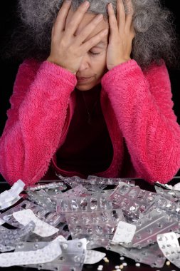 A desperate middle-aged woman with her hands covering her face, is sitting at a table filled with empty medication blisters.