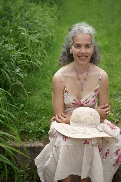 A smiling woman over 50 with curly graying hair is sitting in a garden. Retro Summer dress and straw hat.