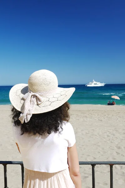 Rear view of a woman with curly hair is wearing a straw hat and is contemplating the sea. In the background, people sitting under an umbrella, a yacht on the horizon.Copy space.