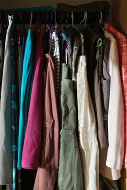 Açık bir gardıroba pantolon, ceket, süveter olarak asılan kadınlara yakın çekim. Otlatıcı güneş ışığı.