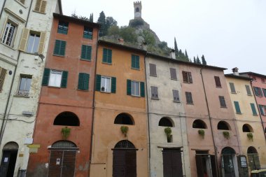 Brisighella 'daki renkli binalar. Yükseltilmiş eşek yolunun kemerli pencereleri ve arka plandaki tepenin üstündeki saat kulesi.