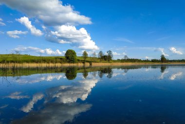 Gölün sakin yüzeyi bir ayna gibi parlak mavi gökyüzünü yansıtır. Baharda doğa yürüyüşü yapan bulutlar.