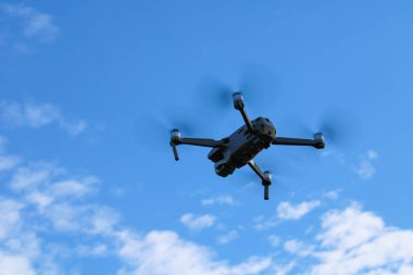 Mavi gökyüzünün arka planında aşağıdan bulutlar görünen kuadkopter dronu uçuyor.