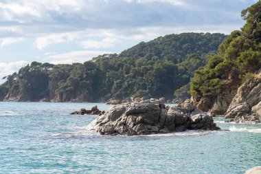 Kayaların etrafında küçük dalgalar ve arka planda yeşillik olan tepeler olan güzel deniz manzarası.