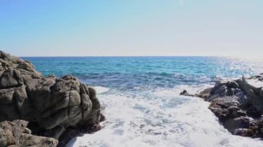 Deniz dalgaları arka planda açık mavi gökyüzü olan kayalara çarpar.