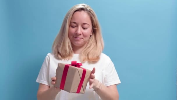 Junge lächelnde Frau hält Geschenkschachtel in der Hand und reicht sie der Kamera auf blauem Grund — Stockvideo