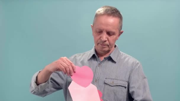 У віці чоловік тримає відкритий рожевий конверт з рожевим паперовим серцем на синьому фоні — стокове відео